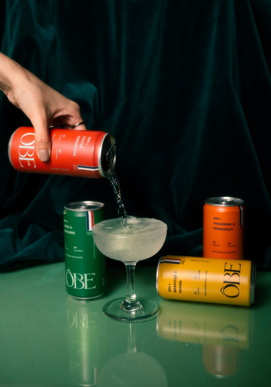 Mão servindo o drink de gin Frutas Vermelhas da Ôbe numa taça, com as latas de Limão & Gengibre, Tangerina & Grapefruit e Maracujá & Manga em volta, sobre fundo verde escuro